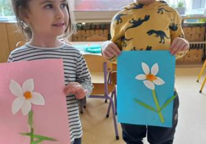 Chłopiec i dziewczynka trzymają w dłoniach zrobione przez siebie prace plastyczne przedstawiające kwiatki wykonane z wacików kosmetycznych, bibuły oraz elementów zielonego papieru kolorowego