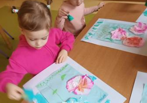 Dwie dziewczynki stemplują farbą przy użyciu gąbki ramkę pracy plastycznej
