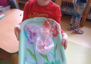 Chłopiec trzyma w dłoniach podkładkę z efektami swoich działań twórczych i przedstawia swoją "Kompozycję kwiatową"