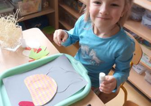 Dziewczynka przy stoliku układa kompozycję z elementów kolorowego papieru przedstawiającą kurę