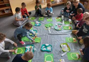 Dzieci siedzą na dywanie i wykonują prace plastyczną zgodnie z instrukcją nauczycielki przedstawiającą koszyczek wielkanocny