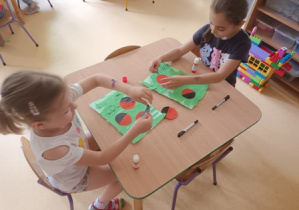 Dziewczynki siedzą przy stoliku w czasie wykonywania pracy plastycznej i naklejają kompozycje biedronek z elementów kolorowego papieru