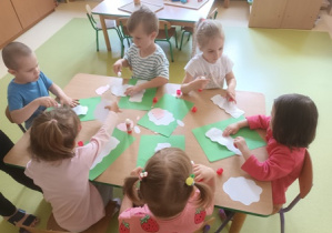 Dzieci młodsze siedzą pry stoliku i naklejają elementy pracy plastycznej na zieloną karkę papieru