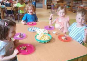 Dzieci w czasie urodzinowego poczęstunku siedzą przy stołach i jedzą drobne słodkości oraz owoce