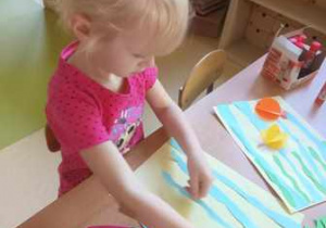 Dziewczynka przykleja na karton wydarte przez siebie paski niebieskiego papieru