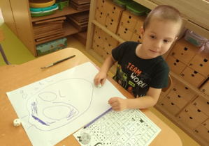Chłopiec siedzi przy stoliku i rysuje "Kropkę" na postawie gry matematycznej "Wylosuj mnie"