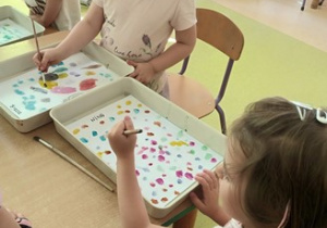 Dwie dziewczynki malują farbami akwarelowymi kolorowe kropki na białym tle