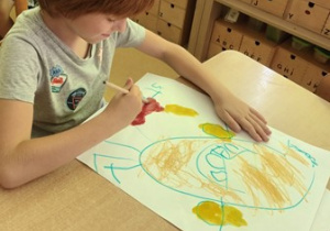 Chłopiec siedzi przy stoliku i maluje tło farbami na swojej pracy plastycznej pt. "Kropka"