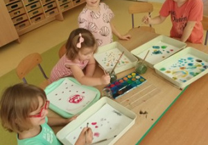 Dzieci młodsze siedzą przy stoliku i malują farbami akwarelowymi kolorowe kropki na białym tle