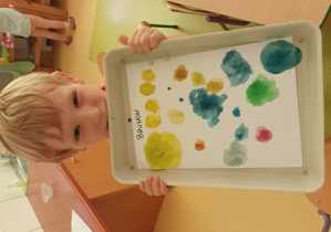 Chłopiec trzyma w dłoniach swoją pracę plastyczną - namalowane kolorowe kropki przy użyciu farb akwarelowych