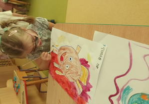 Dziewczynka uzupełnia swoją pracę plastyczną i maluje czerwone tło farbą akwarelową