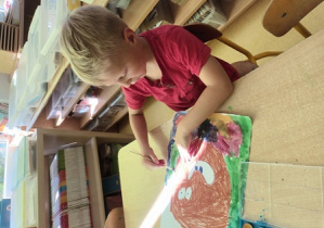 Chłopiec siedzi przy stoliku i maluje farbami tło swojej tematycznej pracy plastycznej "Kropka"