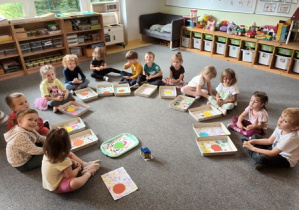 Dzieci młodsze siedzą na dywanie i naklejają elementy kolorowego papieru na wcześniej przygotowane tło w kropki