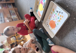 Dziewczynka i chłopiec siedzą na dywanie i odkręcają klej w sztyfcie, przed dziećmi na widać podkładki z tematyczną pracą plastyczną "Kropki"