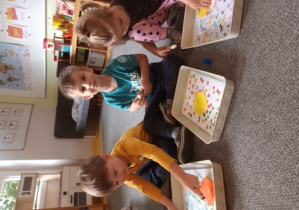 Dzieci siedzą na dywanie i przyklejają kropkę z kolorowego papieru na swoje wykropkowane wcześniej tła