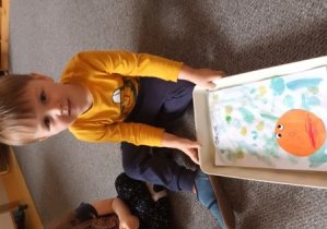 Chłopiec siedzi na dywanie i trzyma w dłoniach swoją pracę plastyczną
