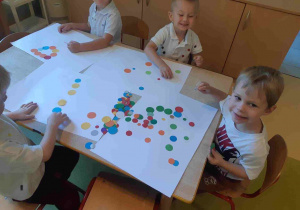 Dzieci siedzą przy stole i przyklejają kropki na karton