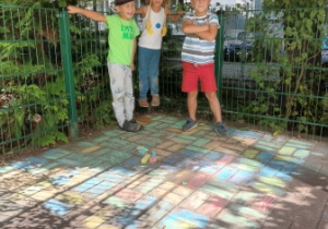 Trzej chłopcy pozują do zdjęcia z namalowaną przez siebie kredami kolorową kompozycją