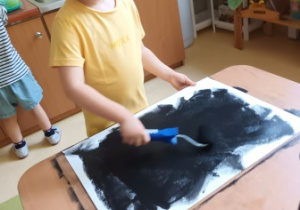 Chłopiec maluje tło do pracy plastycznej na dużym arkuszu przy użyciu wałka