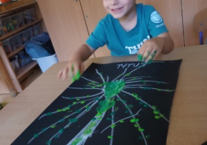 Chłopiec siedzi przy stoliku stempluje palcami umoczonymi w zielonej farbie na swojej pracy plastycznej