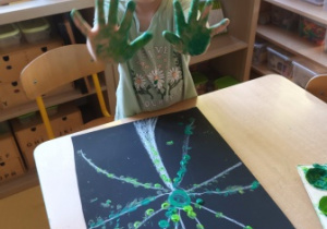 Anielka pozuje do zdjęcia z wykonaną przez siebie pracą plastyczną oraz dłońmi umalowanymi zieloną farbą