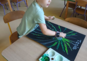 Dziewczynka rozprowadza na swojej pracy plastycznej zieloną farbę poprzez rozcieranie palcami