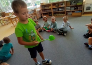 Chłopiec podczas ćwiczenia ciszy ćwiczy równowagę trzymając w ręce zieloną łyżkę z zieloną piłką, w tle widać dzieci siedzące w kręcu
