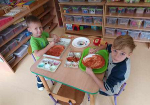 Chłopcy siedzą przy stoliku i malują farbami powierzchnię papierowego talerzyka brązową farbą