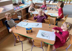Dzieci starsze siedzą przy stoliku i domalowują drugą część portretu Marii Montessori pastelami olejnymi