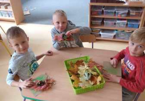 Chłopcy siedzą przy stoliku i układają kompozycję z jesiennych liści i wcześniej wykonanych ślimaków z plasteliny