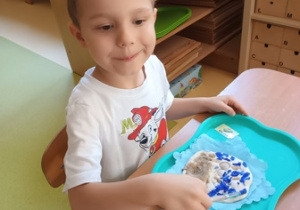 Chłopiec siedzi przy stoliku i uzupełnia swoją kompozycje z masy solnej malując niebieską farbą