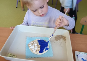 Chłopiec siedzi przy stoliku i rozprowadza pędzlem niebieską farbę na powierzchni masy solnej
