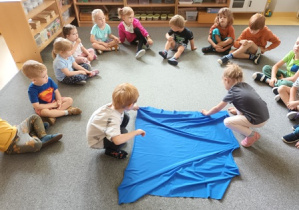 Dzieci siedzą w kręgu na dywanie podczas urodzin, Brunon i Liwia rozkładają niebieski materiał w środku kręgu