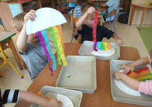 Dzieci w czasie wykonywania pracy plastycznej siedzą przy stoliku i doklejają kolorowe paski bibuły do powierzchni papierowego talerzyka