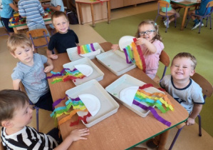 Dzieci siedzą przy stoliku i nazywają poszczególne kolory bibuły doklejonej przez siebie do papierowego talerzyka