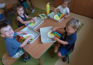 Dzieci młodsze siedzą przy stoliku i wykonują pracę plastyczną stemplując powierzchnie papierowego talerzyka żółtą farbą