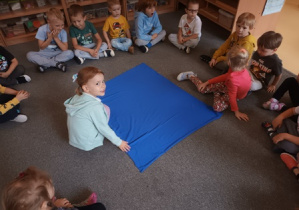 Dzieci siedzą w kręgu na dywanie podczas urodzin, Tosia i Ala rozkładają niebieski materiał w środku kręgu