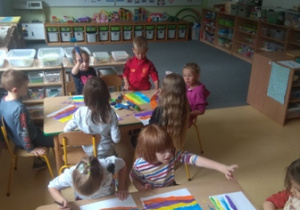 Dzieci siedzą przy stolikach i malują farbami kolorowe paski