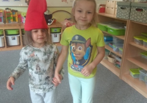 Dwie dziewczynki stoją, jedna ma na głowie czerwoną czapkę, druga uszy królika