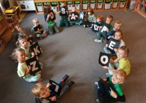 Dzieci siedzą w kręgu i trzymają w rękach różne białe litery umieszczone na czarnym tle