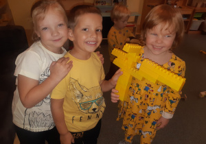 Dwie dziewczynki stoją obok chłopca, który prezentuje budowlę z żółtych klocków