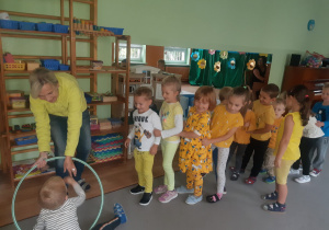 Dzieci ubrane na żółto podczas zabawy ruchowej stoją ustawione w pociągu, jedno za drugim, jedno z dzieci przechodzi przez obręcz trzymaną przez panią