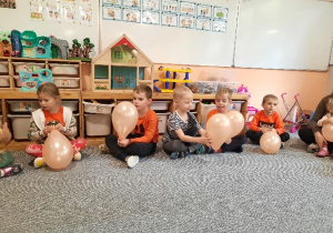 Dzieci ubrane na pomarańczowo siedzą na dywanie i każde z nich trzyma w rękach pomarańczowy balon