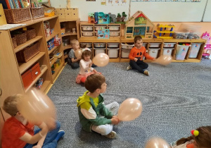 Dzieci ubrane na pomarańczowo siedzą w rozsypce na dywanie i każde z nich trzyma w rękach pomarańczowy balon