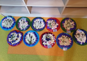 Zdjęcie przedstawia wystawę prac dzieci czyli masę solną rozłożona na kolorowych krążkach, na której umieszczone są kwiaty i fioletowe elementy