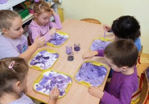 Dzieci siedzą przy stoliku i farbują fioletową farbą kartki umieszczone na tackach