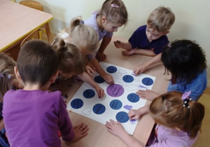 Dzieci siedzą przy stoliku i ozdabiają przyklejone na kartonie fioletowe koła, rysując serduszka