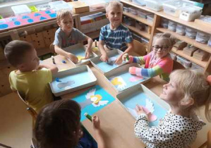 Dzieci starsze siedzą przy stoliku i wykonują pracę plastyczną - układają sylwetę aniołów z elementów kolorowego papieru
