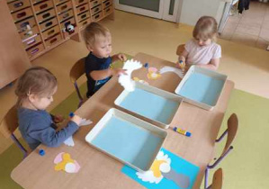 Dzieci młodsze przy stoliku układają kompozycję anioła z elementów kolorowego papieru