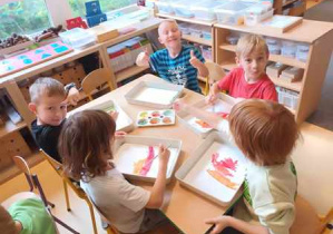 Dzieci starsze siedzą przy stoliku i malują tło pracy plastycznej farbami
